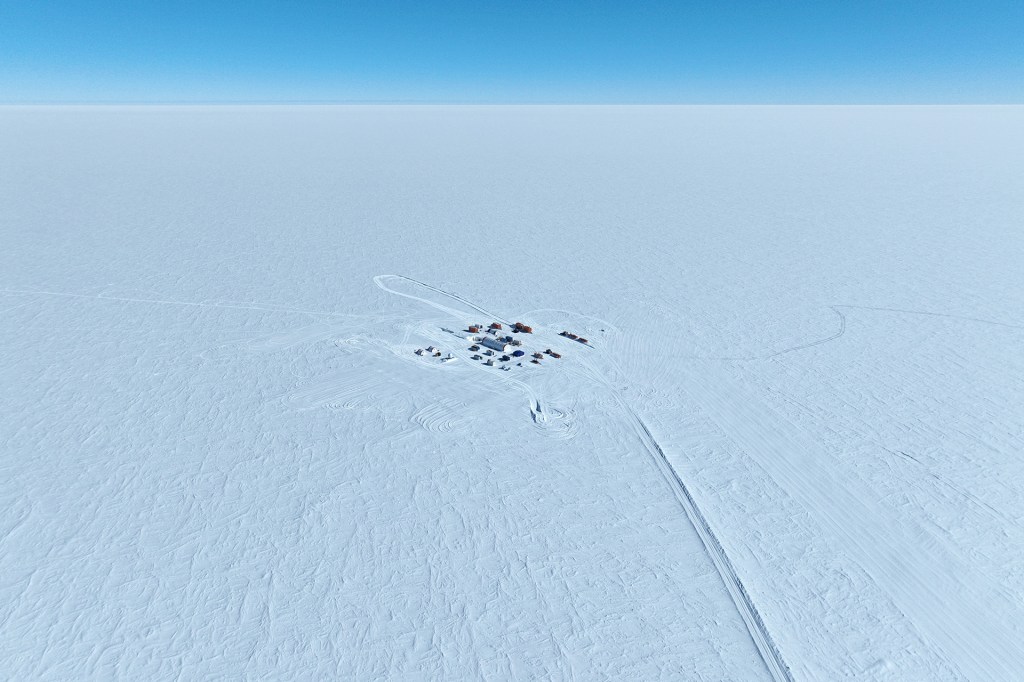 A distant aerial view shows a small research camp alone in a vast, white ice field.