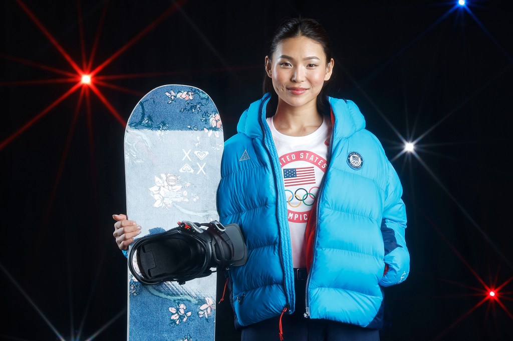 A smiling snowboarder wearing a blue puffer coat and holding a snowboard against a black background.