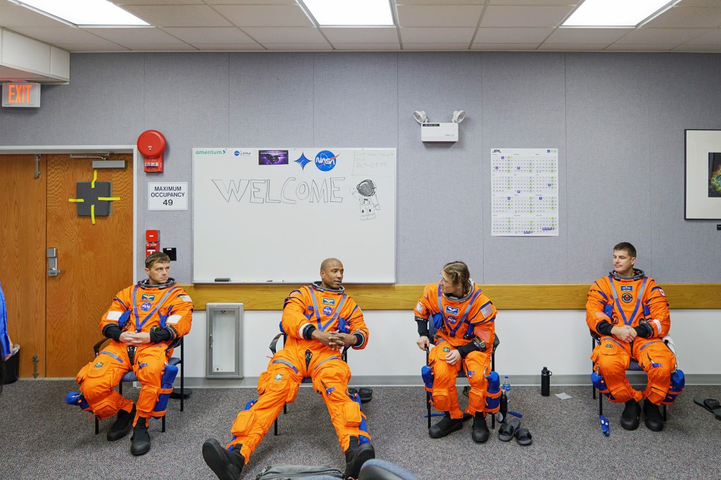 Four astronauts in orange flight suits sit on chairs in a briefing room, talking.