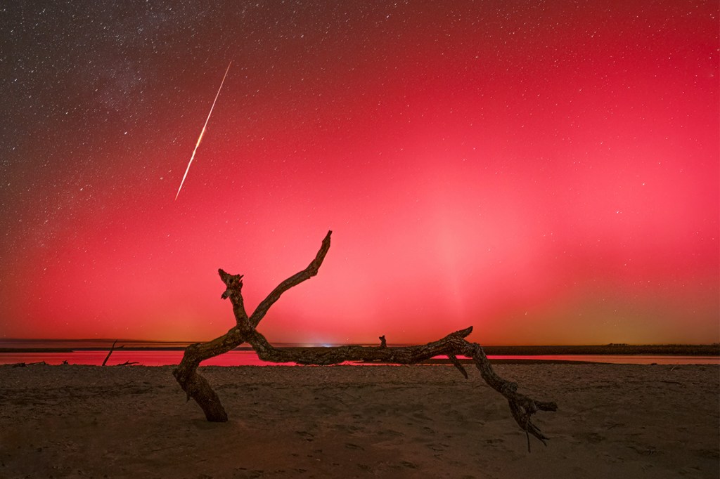 Pink and red northern lights glow across the night sky above a dark shoreline, with a meteor overhead.