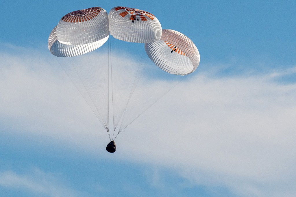 A spacecraft capsule descends through the sky beneath three large parachutes during landing.