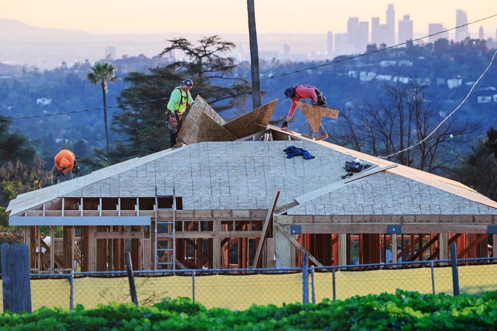 Construction workers repair the roof of a house with a city skyline in the background.