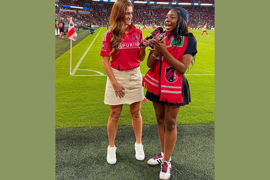 A reporter stands on a soccer field, interviewing a girl wearing a red team scarf.