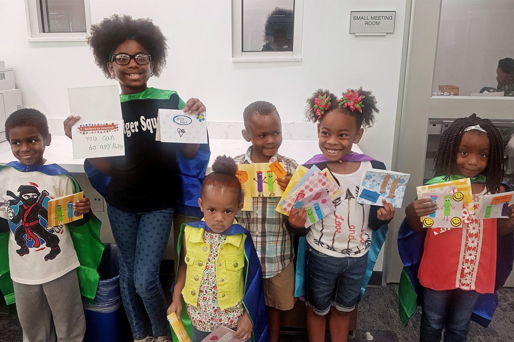 A group of kids wearing capes proudly hold up the cards they made.