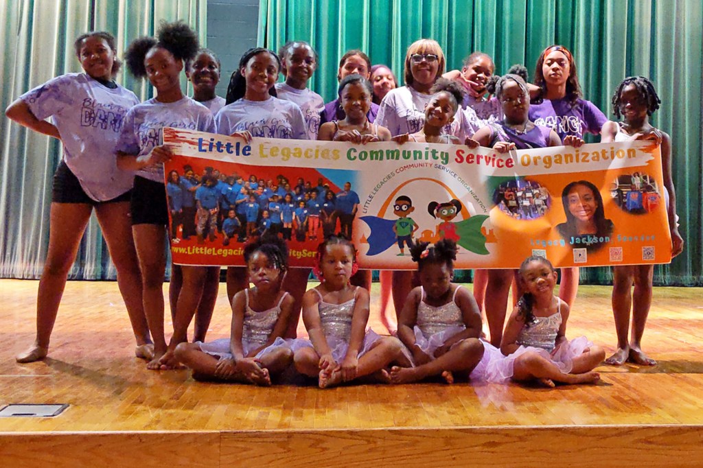 A large group of dancers pose onstage, holding a bright banner for their community group.