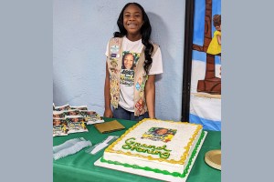 A girl wearing a Girl Scouts vest stands smiling behind a large “Grand Opening” cake displayed on a table at an event.
