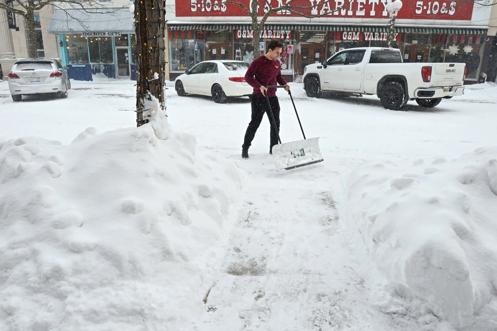 A person uses a wide shovel to clear snow from a sidewalk during a heavy snowfall in a small town.