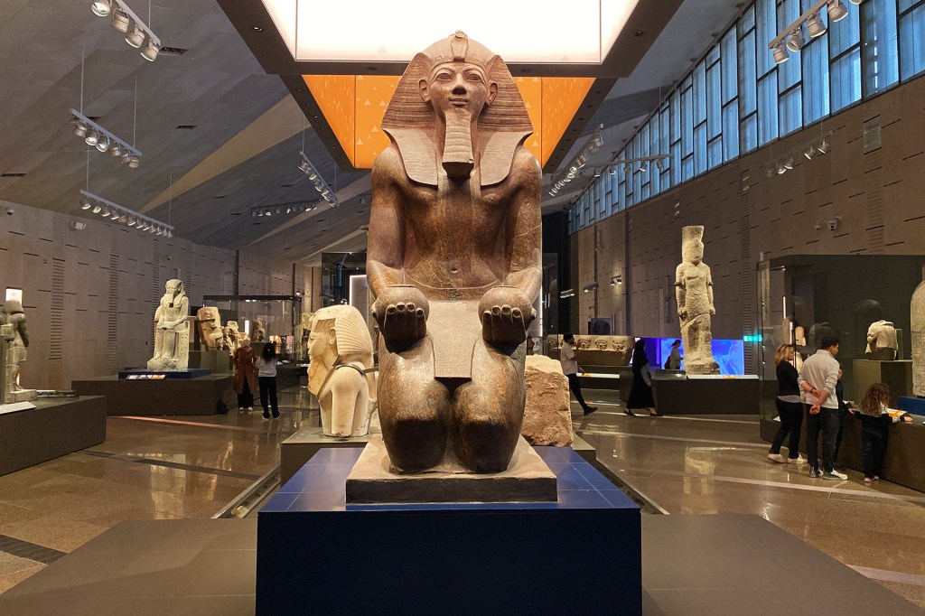 A large stone statue of an ancient Egyptian ruler sits inside a museum, with other statues and visitors in the background.