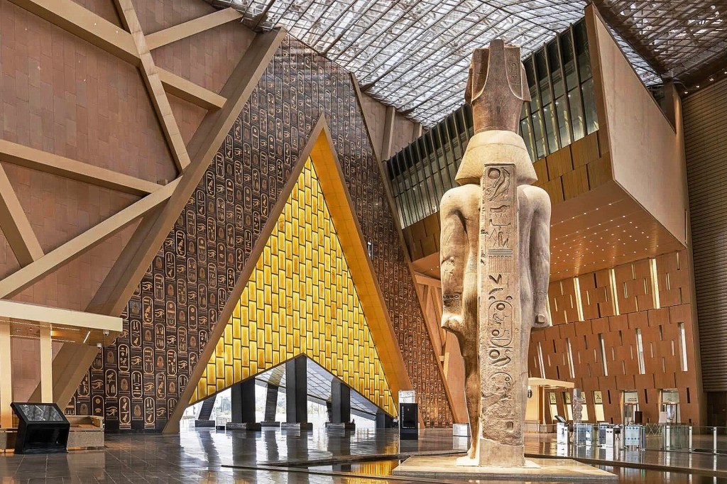 The grand entrance of a museum with a tall statue in the center, high walls, a glass roof, and geometric features of stone and glass.