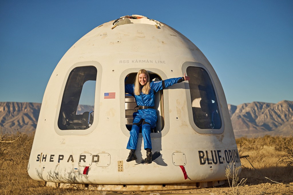 A woman in a blue flight suit sits in the open doorway of a space capsule resting on the ground in a desert.