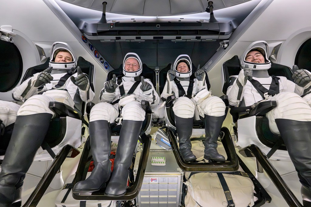 Four astronauts wearing white space suits sit strapped into seats inside a spacecraft cabin.