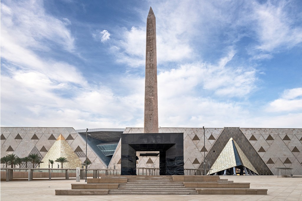 A tall stone obelisk stands outside a museum under a blue sky.