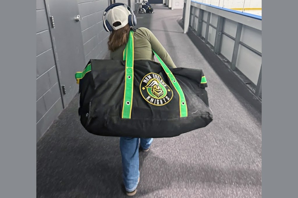 A child wearing headphones walks down a rink hallway carrying a large hockey bag over one shoulder.