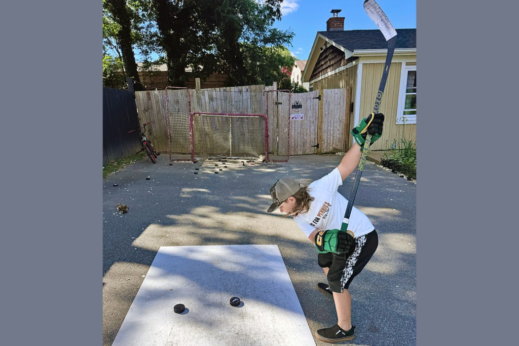 A child practices shooting a hockey puck on a driveway, aiming toward a net set up near a fence.