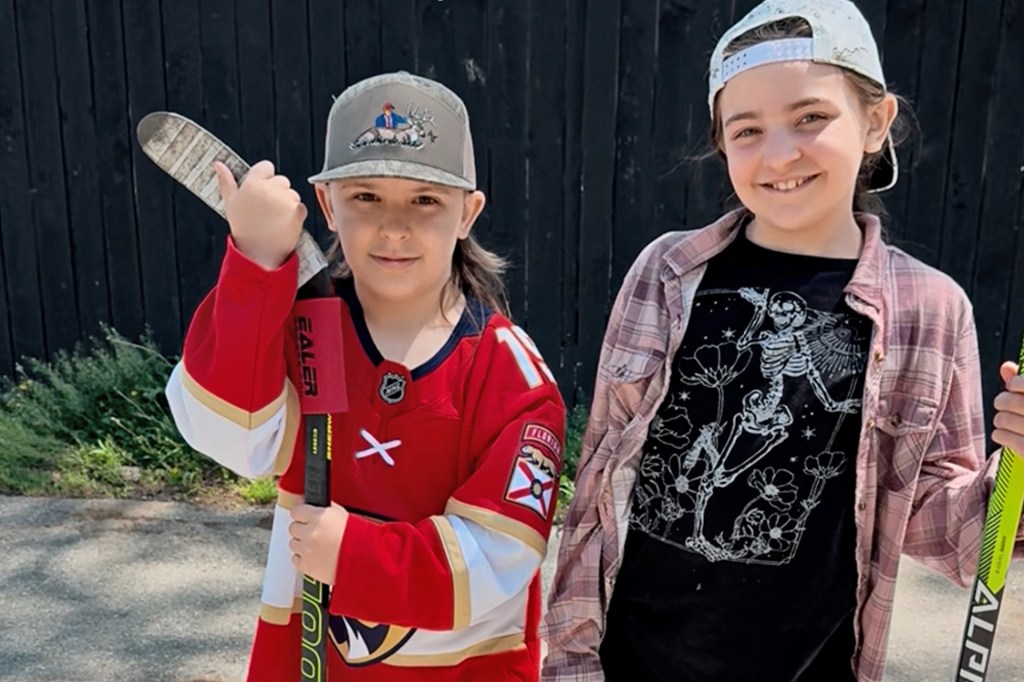 Two children smile while holding hockey sticks, standing side by side outdoors.