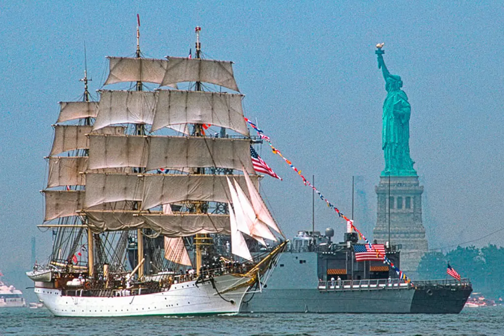 A tall sailing ship passes in front of the Statue of Liberty on the water.