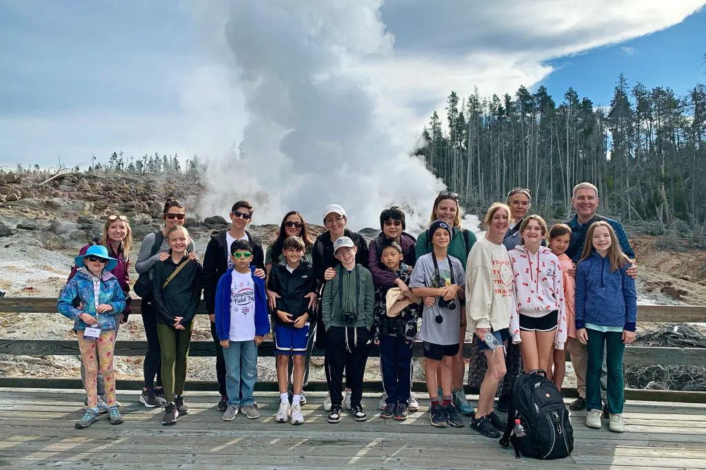 A group of students and adults pose for a photo in front of a steaming geyser in a park.