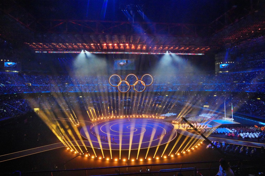 Bright lights shine over a large stadium during an Olympic ceremony, with the Olympic rings glowing above the stage.