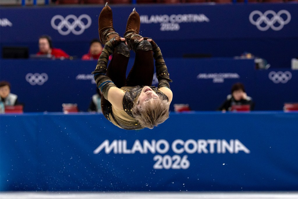 A figure skater flips upside down in midair during a performance, with a sign reading “Milano Cortina 2026” behind.