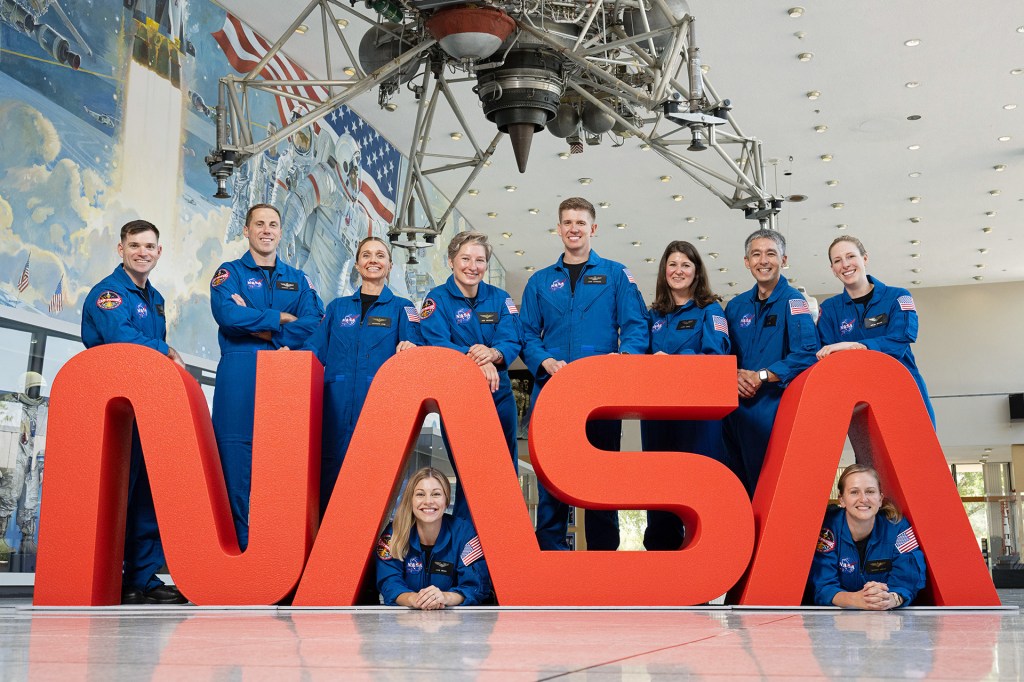 A group of NASA astronauts in blue flight suits pose together behind large red letters spelling “NASA.”