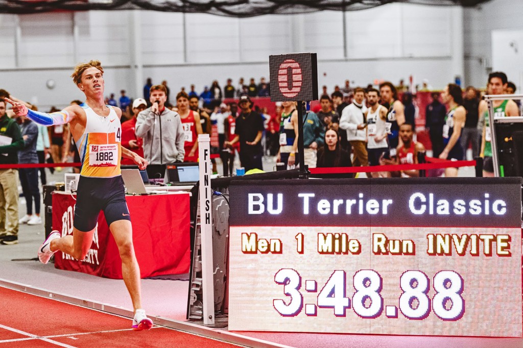 A runner crosses the finish line at an indoor track meet as a digital clock shows the race time.