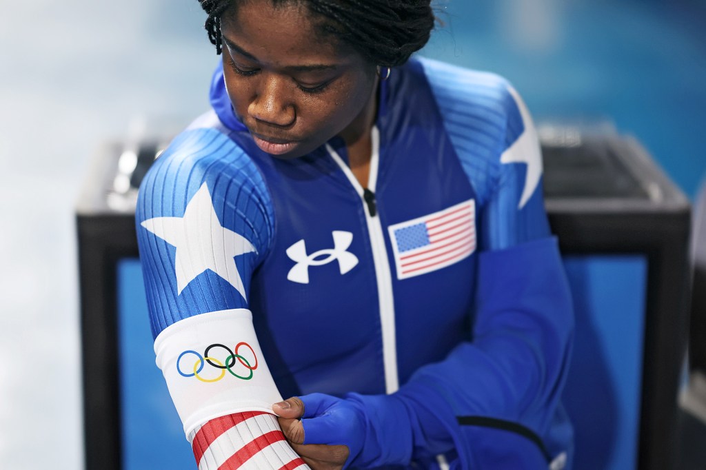 An athlete wearing a blue racing suit adjusts an armband with the Olympic rings and an American flag.