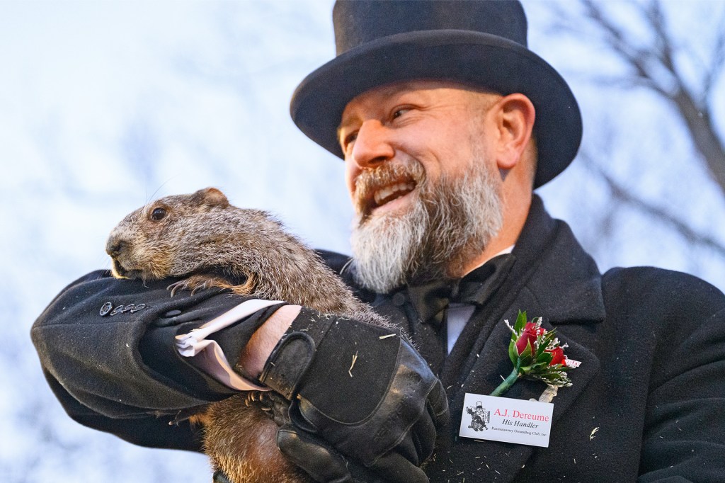 A man wearing a black hat and coat smiles while holding a groundhog during a Groundhog Day event.