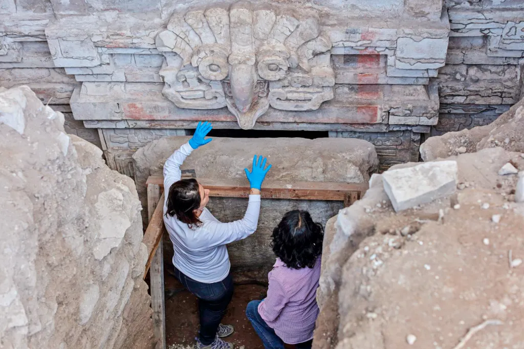 Two archaeologists wearing gloves examine a carved stone wall inside an ancient tomb surrounded by excavation stones.