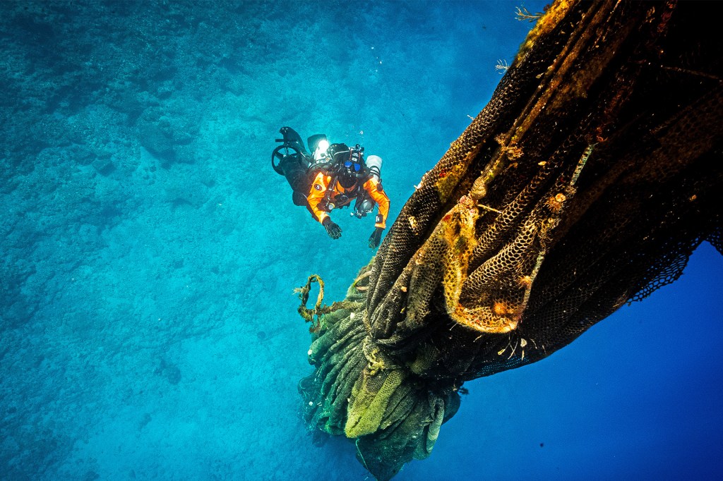 A scuba diver in an orange suit swims underwater beside a large bundle of tangled fishing nets.