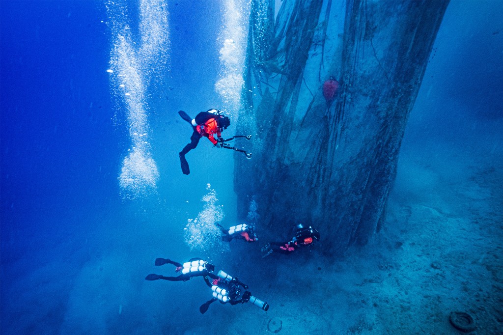 Scuba divers swim underwater beside a large, net-covered structure on the seafloor.