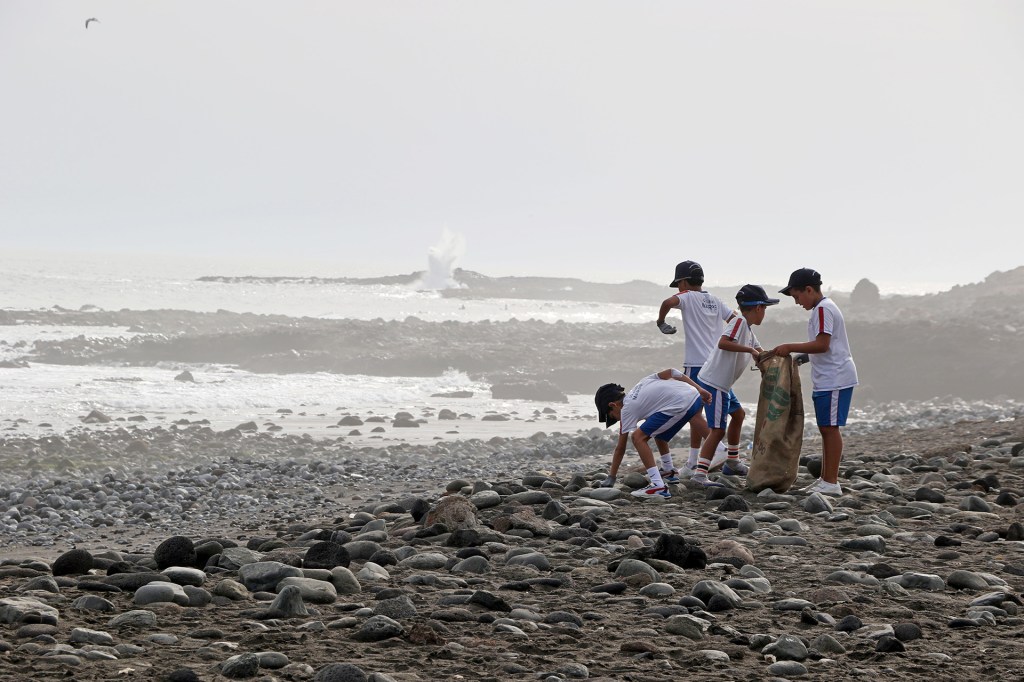 Children pick up trash along a rocky beach and place it into a large bag.