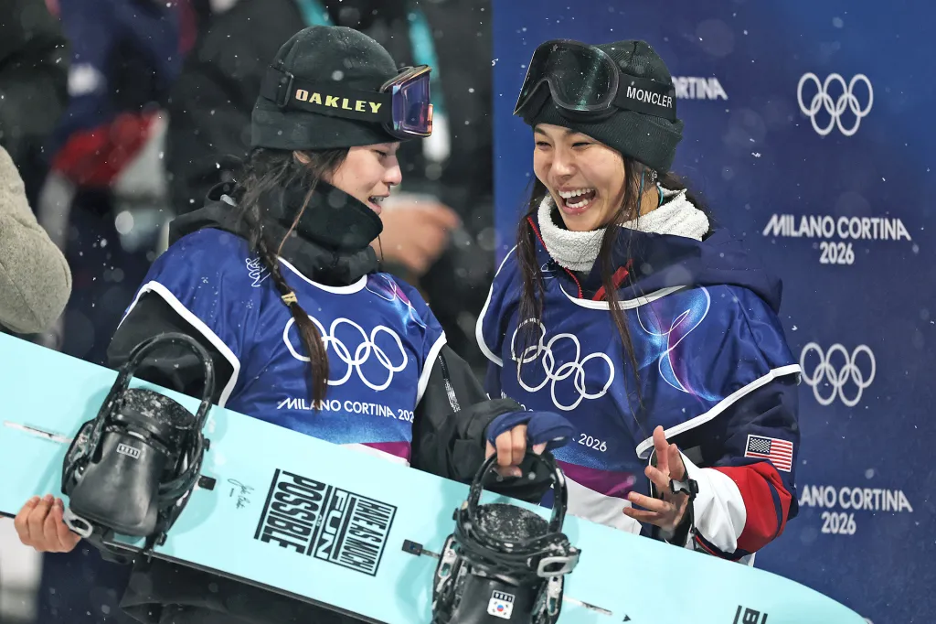 Two snowboarders wearing competition bibs smile and celebrate while holding a snowboard.