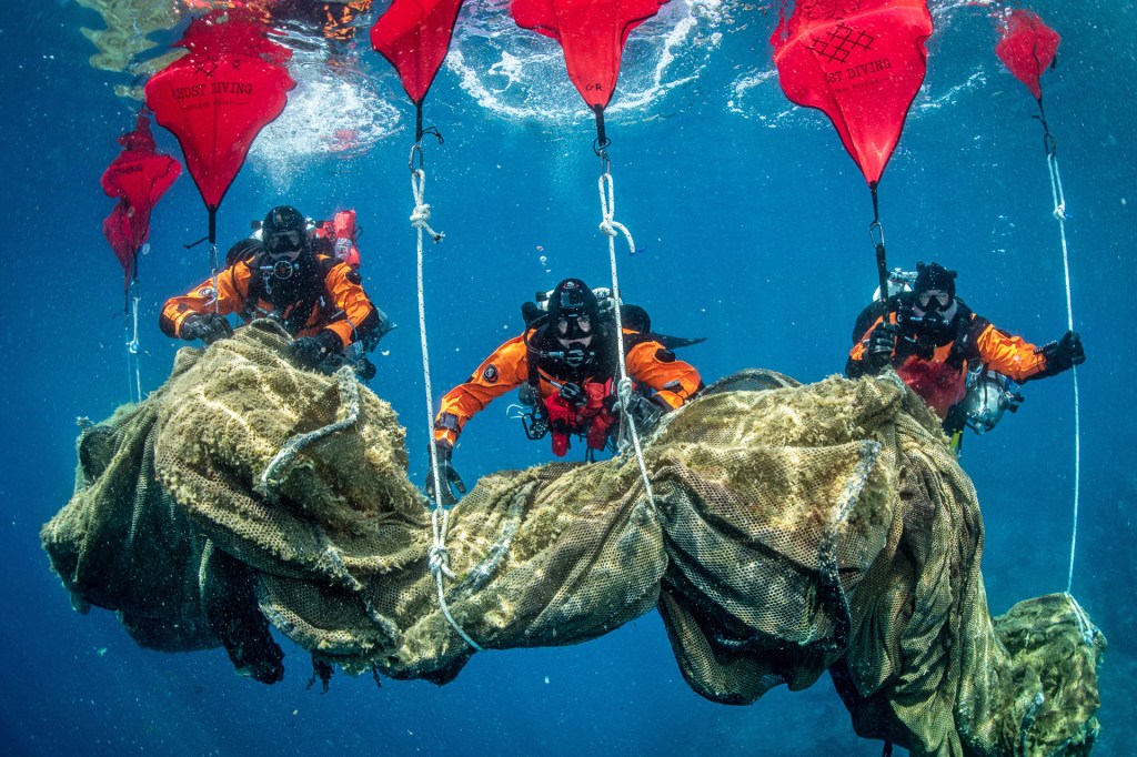 Divers in bright orange suits lift a heavy bundle of netting underwater using floating markers.