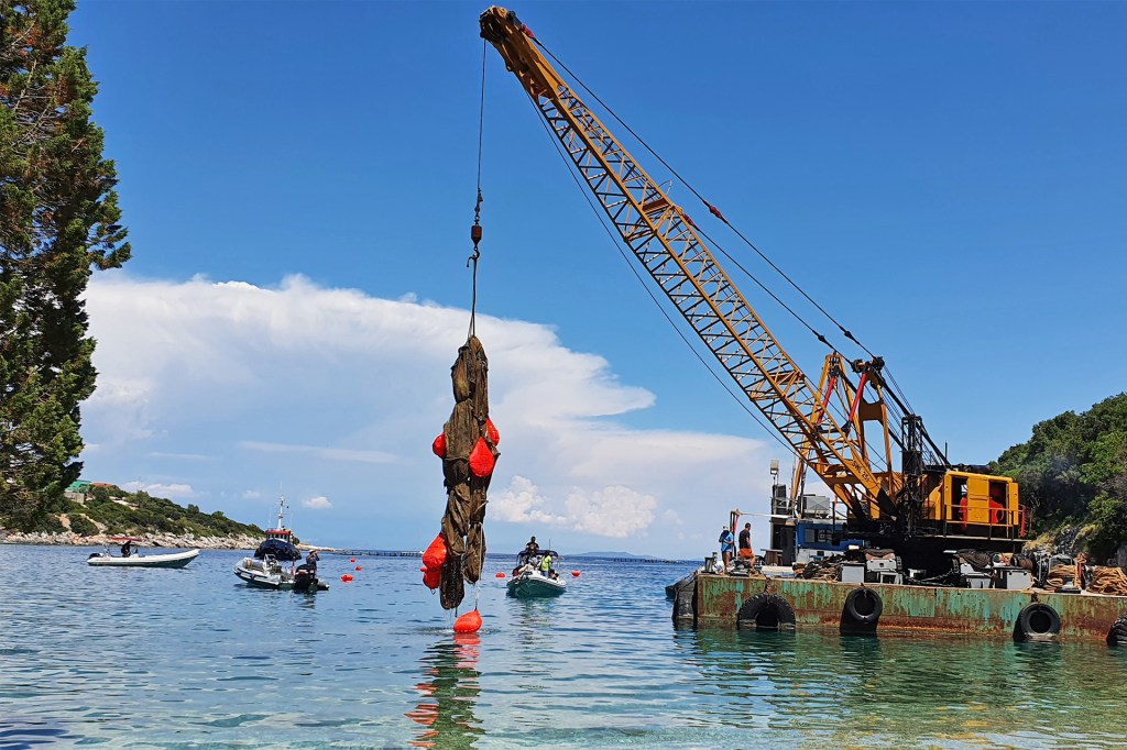 A crane on a barge lifts a large bundle of netting from the water as boats float nearby.