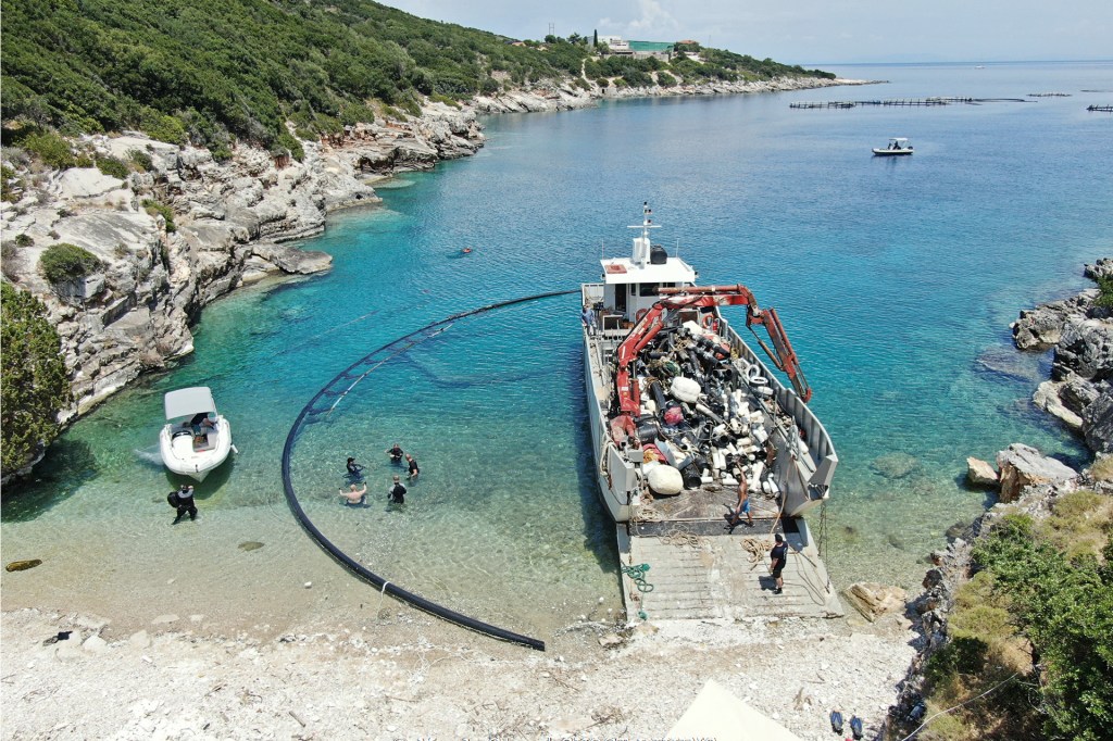A boat filled with collected debris rests in clear blue water near a rocky shoreline.