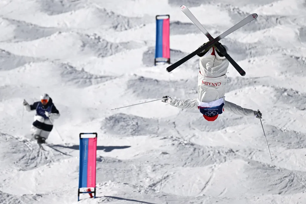 A freestyle skier flips upside down in midair over a snowy course during a competition.