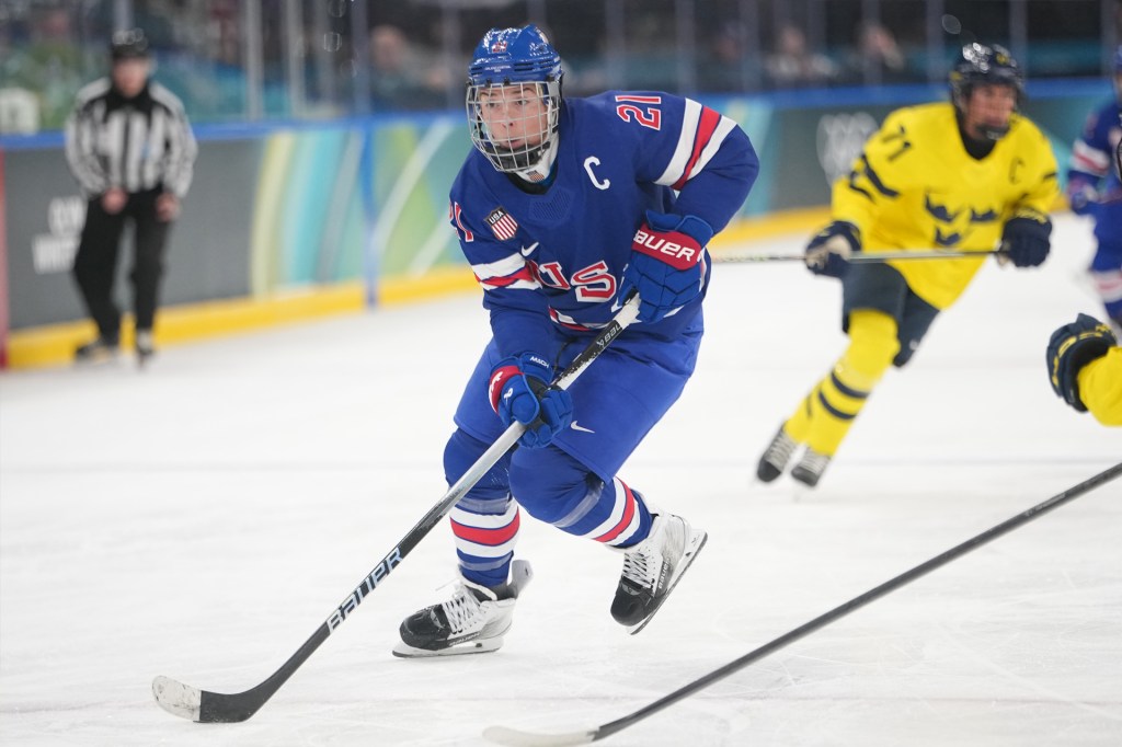 A hockey player in a blue USA uniform skates quickly across the ice while controlling the puck, with a player in a yellow uniform chasing behind.