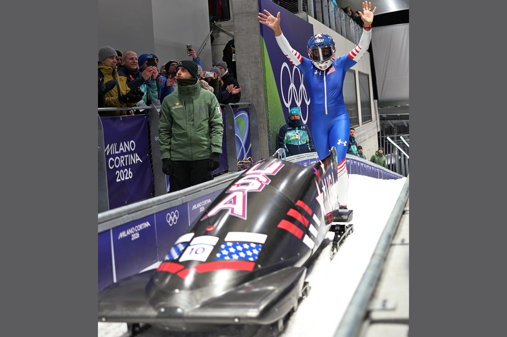 A bobsledder in a blue racing suit raises her arms at the finish line beside her sled as fans cheer.