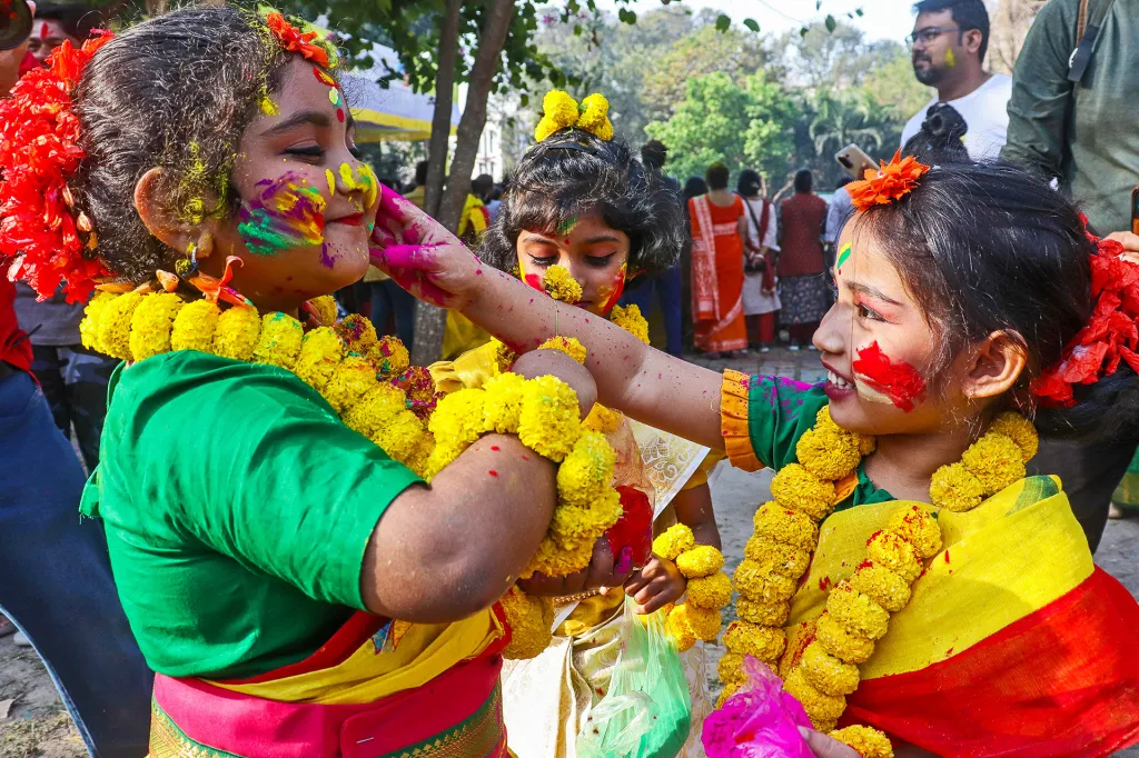 Two girls wearing bright flowers smile and put colorful powder on each other’s faces during a festival.