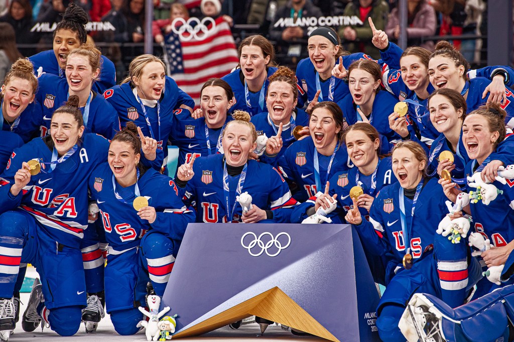 The U.S. women’s hockey team poses together on the ice, smiling and holding up their gold medals.