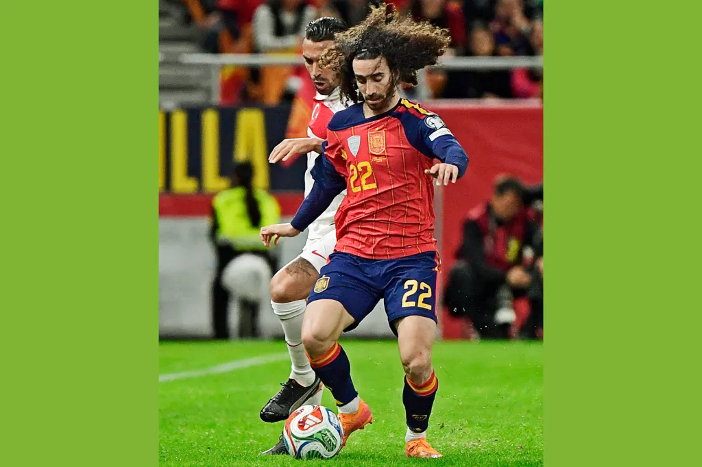 A soccer player in a red uniform dribbles the ball while another player follows behind.