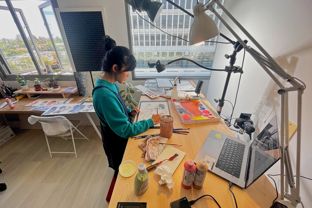 An artist sits at a desk by a window, painting and drawing while art supplies and sketches are spread out around her.