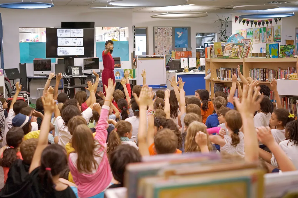 A person stands at the front of a classroom, showing drawings while many students raise their hands.