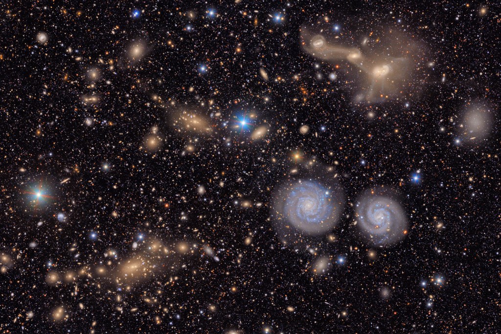A deep-space image filled with thousands of stars and many galaxies, including two bright blue spiral galaxies near the center.