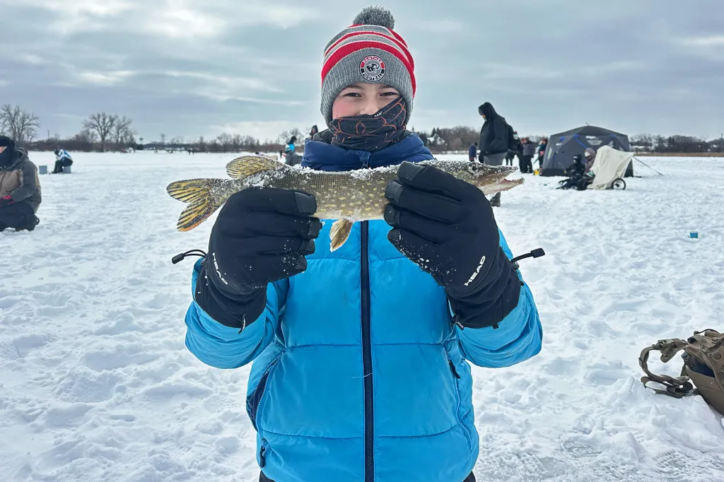 A smiling boy wearing a blue coat holds a fish. He's standing in a snowy environment with a tent in the background.