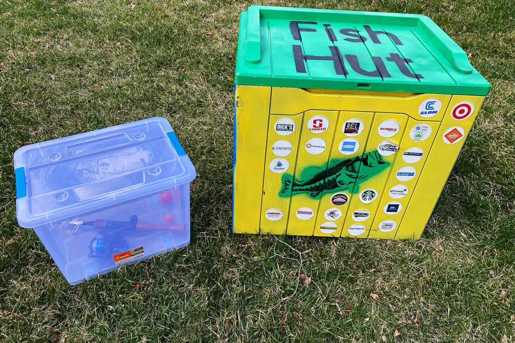 A clear plastic bin containing a fishing rod next to a wooden box with the words "Fish Hut" sitting in the grass.