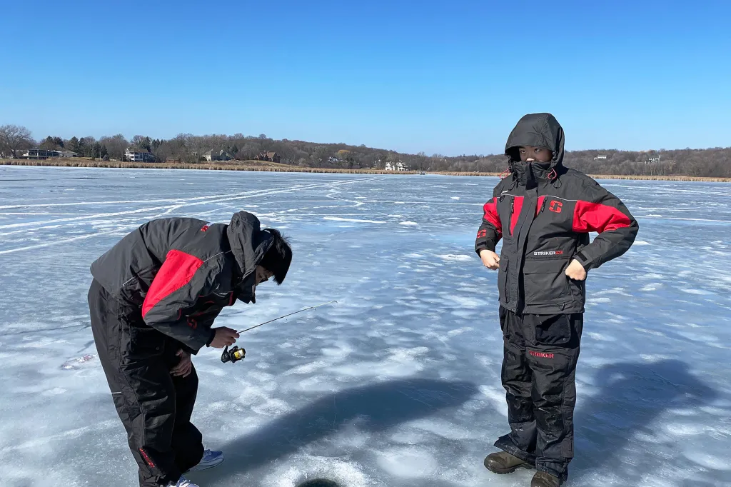 Two kids stand on a frozen lake wearing winter gear. One holds a fishing rod and leans forward to look in a hole drilled in the ice.