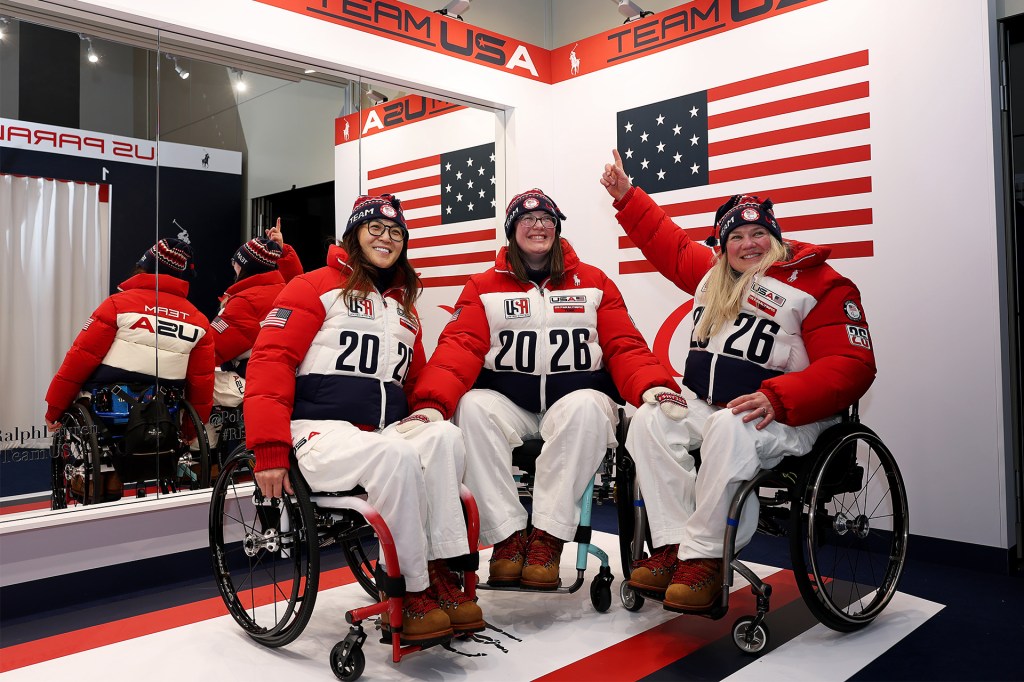 Three athletes in wheelchairs wear red, white, and blue Team USA jackets and smile in front of large American flags.