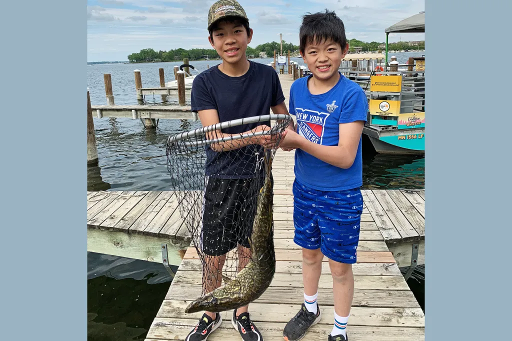 Two smiling boys stand on a dock, holding a fishing net containing a large fish.