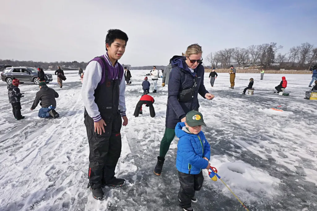 A boy wearing snow pants stands near a younger child holding a fishing rod and his mom on a frozen lake.